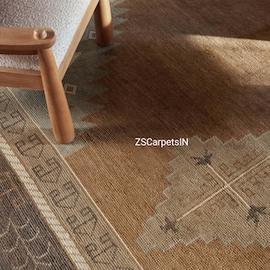 May include: A close-up of a brown and beige patterned rug with a geometric design. The rug has a border with a repeating pattern and is partially covered by a wooden chair. The text "ZSCarpetsIN" is visible on the rug.