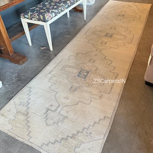 May include: A long, rectangular beige rug with a geometric pattern in shades of grey. The rug features a central design with repeating shapes and a border with a zigzag pattern. The text "ZSCarpetsIN" is visible on the rug.