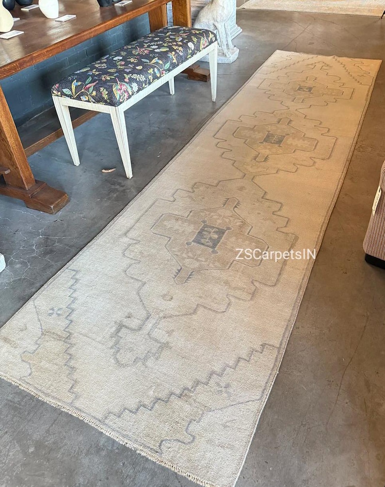 May include: A long, rectangular beige rug with a geometric pattern in shades of grey. The rug features a central design with repeating shapes and a border with a zigzag pattern. The text "ZSCarpetsIN" is visible on the rug.