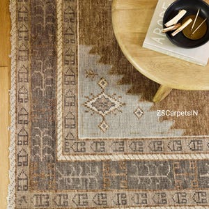 May include: A rectangular area rug with a geometric pattern in shades of brown, beige, and light blue. The rug has a border with repeating geometric shapes and a fringed edge. A wooden table and a book are in the background.