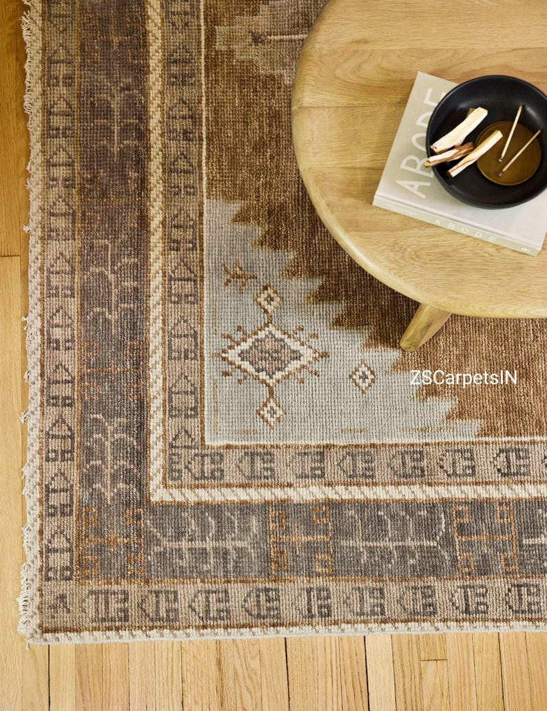 May include: A rectangular area rug with a geometric pattern in shades of brown, beige, and light blue. The rug has a border with repeating geometric shapes and a fringed edge. A wooden table and a book are in the background.