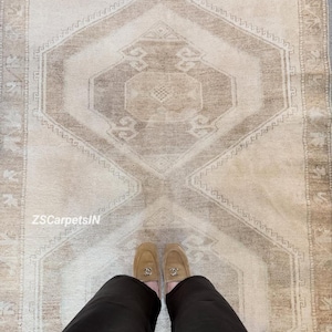 Op de afbeelding: Beige en bruin patroonkleed met een geometrisch ontwerp. Het kleed heeft een centraal medaillon en borderdetails. De tekst "ZSCarpetsIN" is zichtbaar. De afbeelding toont ook de benen en voeten van een persoon die beige loafers draagt.
