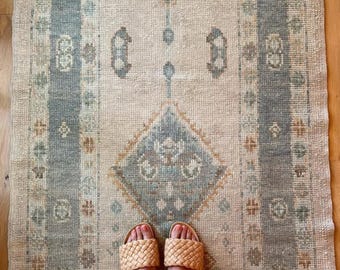 May include: A rectangular rug with a beige background and geometric patterns in shades of blue, green, and gold. The rug has a central diamond design and borders with vertical stripes. Woven sandals are visible at the bottom.