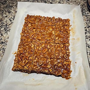 Op de afbeelding: Pecannoten taartreepjes gesneden in vierkante porties. De repen liggen op bakpapier. De bovenste laag is een glanzend, karamelkleurig glazuur, bezaaid met pecannoten. De repen zijn goudbruin van kleur.