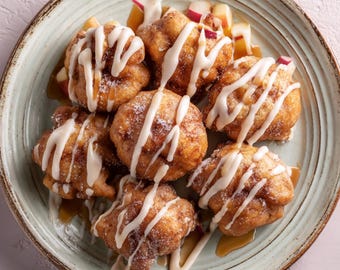 May include: A plate of golden-brown, sugared apple fritters drizzled with white icing and a caramel sauce. The fritters are arranged on a light green plate, garnished with diced apples. A delicious dessert.
