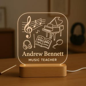 May include: A personalised music-themed LED night light. The clear acrylic panel features musical notes, a piano, headphones, and sheet music. The name "Andrew Bennett" and the text "MUSIC TEACHER" are displayed. The light sits on a wooden base.