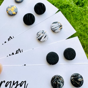 May include: A collection of handmade stud earrings displayed on white cards. The earrings feature various designs, including black textured, marble-effect with gold accents, and gray with gold leaf. The "Raya Craft Studio" logo is visible.