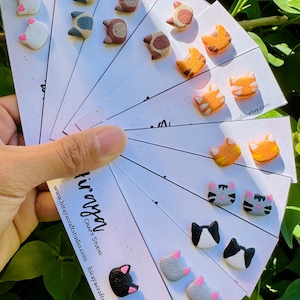 May include: A variety of cat-shaped stud earrings in different colors, including white, gray, black, orange, and brown. The earrings are displayed on small white cards. The cards have the text "hiraya CRAFT STUDIO" and a website address.