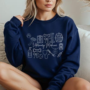 May include: Navy blue sweatshirt with white line art illustrations of veterinary tools and pet-related items. The words "Veterinary Medicine" are written in cursive. The design includes a paw print, brush, bone, cat carrier, and more.