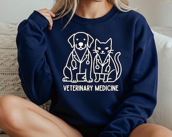 May include: Navy blue sweatshirt featuring a white graphic of a dog and cat wearing lab coats and stethoscopes. The text "VETERINARY MEDICINE" is printed below the illustration. The sweatshirt is a crew neck style.