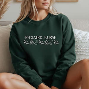 May include: A dark green sweatshirt featuring the white text "PEDIATRIC NURSE". Below the text are white illustrations of a stethoscope, flower, dinosaur, flower, dinosaur, and stethoscope. The sweatshirt has a crew neck.