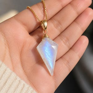 Può includere: Un ciondolo in pietra di luna sfaccettata a forma di aquilone con una distinta adularescenza blu, sospeso a una sottile catena a scatola dorata. La gemma è fissata da una montatura in filo metallico dorato, mostrando un aspetto bianco traslucido.
