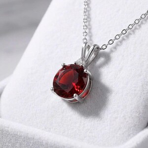 May include: A silver necklace featuring a large, oval-cut, deep red gemstone pendant. The pendant is set in a silver bezel with a small accent stone. The chain is delicate and silver, creating an elegant look.