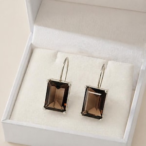 May include: A pair of rectangular smoky quartz earrings with silver hooks, presented in a white jewellery box. The earrings feature a dark brown, transparent appearance, set in silver-toned metal.