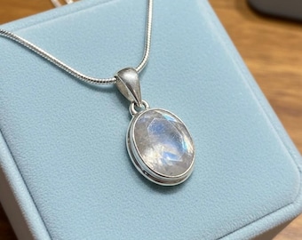 May include: A silver necklace with an oval moonstone pendant. The pendant is set in a silver bezel and has a shimmering, iridescent quality. The necklace is displayed on a light blue jewelry box.