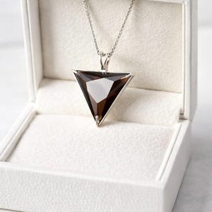 Op de afbeelding: Een zilveren ketting met een driehoekige hanger met een facetgeslepen, rookbruine edelsteen. De hanger wordt getoond in een open witte sieradendoos met een zacht, gestructureerd interieur. De ketting is delicaat.