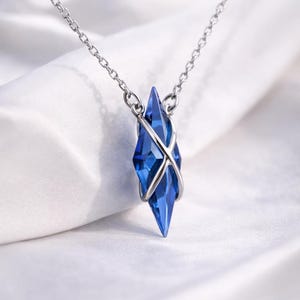 May include: A silver necklace featuring a faceted, blue crystal pendant. The crystal is diamond-shaped and encased in a silver metal frame. The pendant hangs from a delicate silver chain, creating a simple yet elegant design.