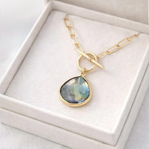 Op de afbeelding: Goudkleurige ketting met een druppelvormige hanger. De hanger heeft een veelkleurige steen met blauwe, groene en grijze tinten, gezet in een gouden frame. De ketting heeft een tuimelsluiting en wordt gepresenteerd in een witte sieradendoos.