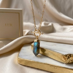 May include: A gold-toned necklace featuring a long, faceted labradorite pendant with blue iridescence, suspended from a gold chain with a toggle clasp. The toggle bar is accented with two small, clear stones on each end, and the chain links are elongated.