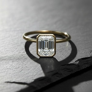May include: A gold ring featuring an emerald-cut diamond. The diamond is set in a bezel setting, and the band is thin and polished. The ring is displayed on a dark surface, casting a shadow.