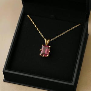 Könnte beinhalten: Eine goldene Halskette mit einem rechteckigen, rubinroten Anhänger, präsentiert in einer schwarzen Schmuckschatulle. Der Anhänger hat einen tiefroten Farbton, und die Kette ist zart. Die Schatulle ist geöffnet und zeigt die Halskette vor schwarzem Hintergrund.