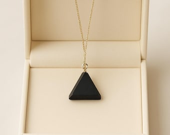 Puede incluir: Un collar con colgante triangular negro y cadena dorada, presentado en un joyero crema abierto. El colgante tiene una forma geométrica. El collar es una joya sencilla y elegante.