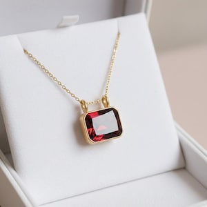 May include: A gold necklace with a rectangular, deep red gemstone pendant. The pendant is set in a gold frame and hangs from a delicate gold chain. The necklace is displayed in a white jewelry box.