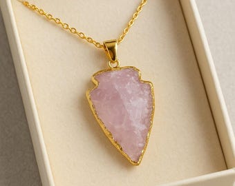Puede incluir: Un collar dorado con un colgante en forma de punta de flecha. El colgante es una piedra rosa claro, tallada en bruto, con un borde dorado. El collar se presenta en una caja color crema.