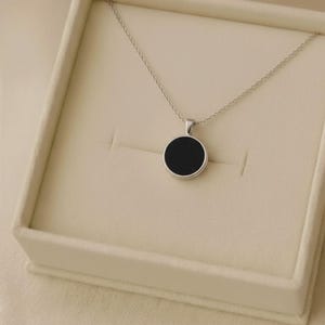 Puede incluir: Un collar plateado con un colgante circular negro, presentado en una caja de joyería color crema. El colgante es un círculo negro sólido, y la cadena es delicada. La caja está abierta, revelando el collar.
