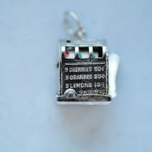 May include: A silver-colored slot machine charm with a loop for a necklace or bracelet. The charm has a detailed design with text that reads "3 Cherries 30-1, 3 Oranges 20-1, 3 Lemons 10-1". The slot machine's reels show cherries, oranges, and lemons.