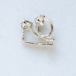 May include: A silver-colored charm in the shape of a swing set with a ring and a floral design. The charm is made of a shiny metal and has a detailed texture. The swing set has a rectangular base and a curved top with a ring for attachment.