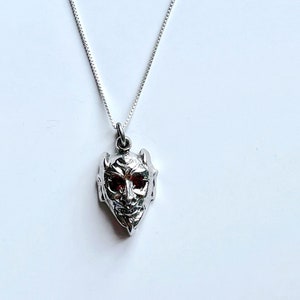 Puede incluir: Un collar de plata con un colgante detallado de la cara de un demonio con ojos rojos.