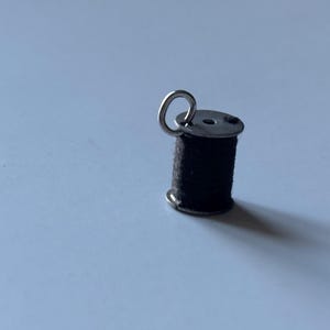 May include: A small metal sewing bobbin charm wrapped in black thread. The cylindrical bobbin features a silver-toned metal top and bottom flange with a circular jump ring attached to the top, allowing it to be used as a pendant or jewelry component.