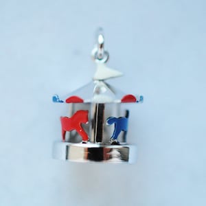 Puede incluir: Un peque&ntilde;o dije de carrusel plateado con caballos rojos y azules. El carrusel tiene un techo plateado con detalles rojos y azules y una peque&ntilde;a bandera blanca. El dije est&aacute; dise&ntilde;ado para ser un colgante o un dije de pulsera.