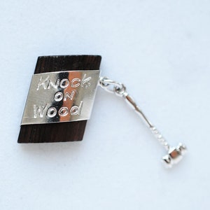 Może przedstawiać: Mały drewniany amulet z srebrną płytką z napisem "Knock on Wood" i małym srebrnym młotkiem przymocowanym do łańcuszka.