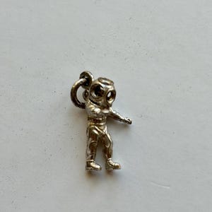 May include: A small, silver-colored charm in the shape of a vintage diving suit. The diver has a round helmet with visible eye holes, a detailed suit, and outstretched arms. The charm includes a loop for attachment, suggesting it's designed for a necklace or bracelet.