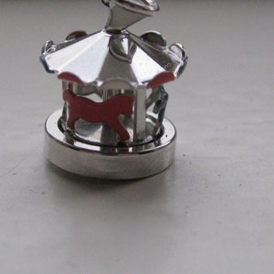 Puede incluir: Un charm de carrusel plateado con un caballo rojo y detalles azules. El charm tiene un dise&ntilde;o detallado con un techo y una base circular. El charm de carrusel es una versi&oacute;n en miniatura de una atracci&oacute;n de feria cl&aacute;sica.