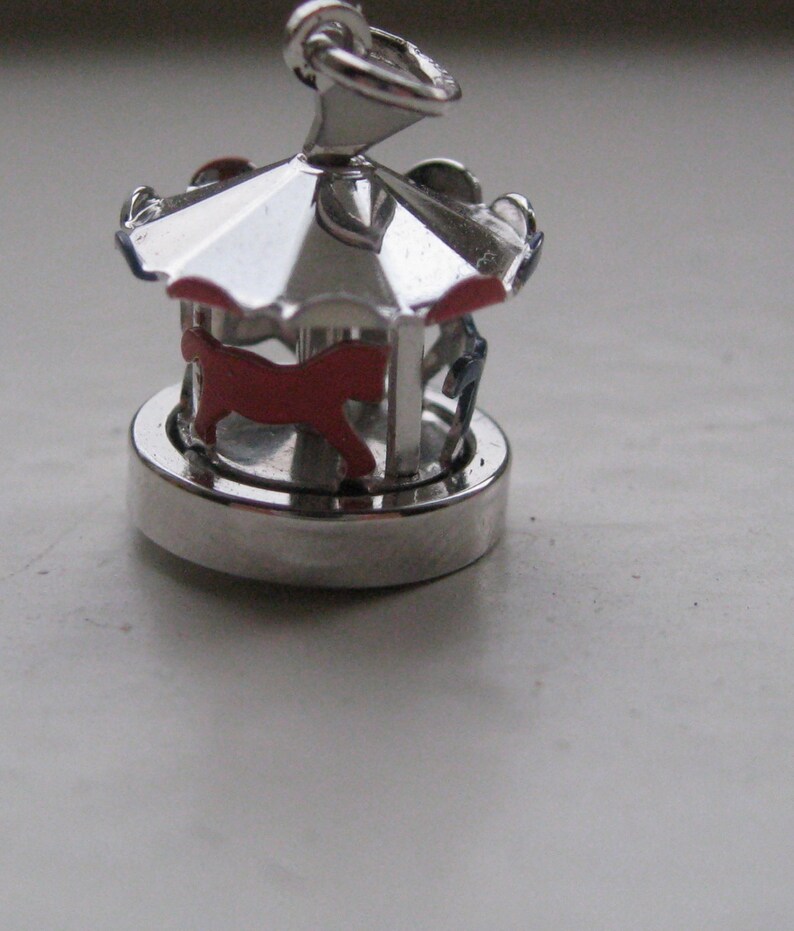 Puede incluir: Un charm de carrusel plateado con un caballo rojo y detalles azules. El charm tiene un dise&ntilde;o detallado con un techo y una base circular. El charm de carrusel es una versi&oacute;n en miniatura de una atracci&oacute;n de feria cl&aacute;sica.