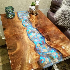Peut inclure: Table d'appoint en bois faite à la main avec un design en forme de rivière. Le centre présente une incrustation de résine bleue remplie de pierres colorées, créant un effet visuel unique. La table mesure environ 76 cm de long et 46 cm de large.