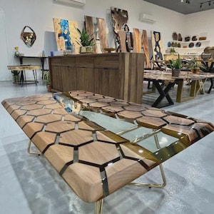 Op de afbeelding: Een rechthoekige houten tafel met een honingraatontwerp, met lichte en donkere houten secties en een transparante harsafwerking. De tafel heeft goudkleurige metalen poten. Andere houten meubels en kunstwerken zijn zichtbaar op de achtergrond.