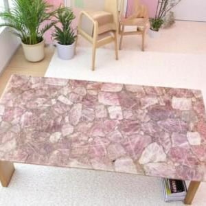 Puede incluir: Una mesa de centro rectangular con una tapa de cuarzo rosa. La superficie de la mesa está compuesta por piedras irregulares de color rosa claro y blanco. La mesa tiene un marco y patas de madera clara. La mesa está en una habitación con plantas y sillas.