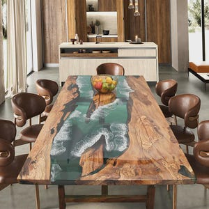 May include: A large, handcrafted wooden dining table with a unique epoxy resin design resembling ocean waves. The table is surrounded by brown leather chairs. A glass bowl of fruit sits on the table. The room has a modern design with wooden cabinetry and a neutral color palette.