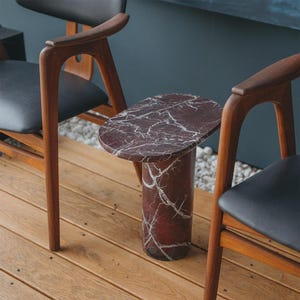 May include: A small, oval-topped side table with a burgundy marble finish and white veining. The cylindrical base complements the tabletop. The table is set on a wooden deck, flanked by two wooden chairs with black seats.