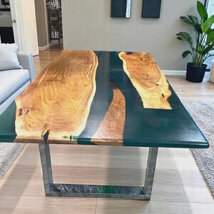 May include: A rectangular coffee table with a dark teal epoxy resin surface and natural wood grain accents. The table has a silver metal base and is in a living room setting. The wood grain is a light brown color.
