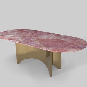 May include: An oval-shaped table with a pink, marbled top and a gold-colored base. The table's base has a unique, curved design. The table is a piece of furniture.