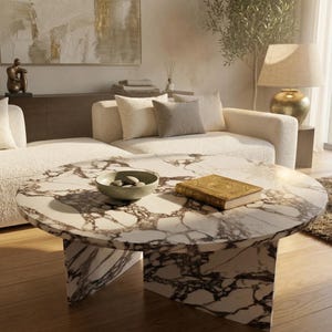 May include: A circular coffee table with a marble top, displaying brown veining. The table has a solid, geometric base. A small bowl with stones and a gold-covered book are on the table. A cream sofa and decorative items are in the background.