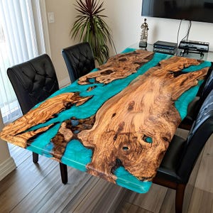 May include: A handcrafted wooden dining table with a turquoise epoxy resin river. The table features a natural wood grain pattern and is supported by dark wooden legs. The table is surrounded by black leather chairs.