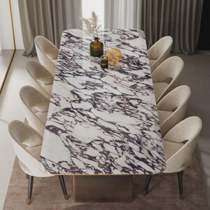 Op de afbeelding: Een rechthoekige eettafel met een wit marmeren blad met donkerpaarse adering. Acht beige gestoffeerde stoelen omringen de tafel. Decoratieve vazen en een kleine sculptuur staan op de tafel. De tafel staat op een tapijt in neutrale tinten.