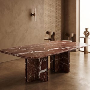 Puede incluir: Una gran mesa de comedor rectangular de mármol rojo con vetas blancas. La mesa tiene un borde redondeado y está sostenida por cuatro patas rectangulares gruesas. La habitación tiene una paleta de colores neutros con una pared de ladrillo y dos sillas.
