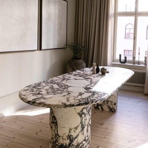 Op de afbeelding: Een grote ovale marmeren eettafel met een wit en zwart geaderd patroon. De tafel heeft een dik, afgerond blad en twee cilindrische voetstukken. De tafel staat in een kamer met hardhouten vloeren en grote ramen.